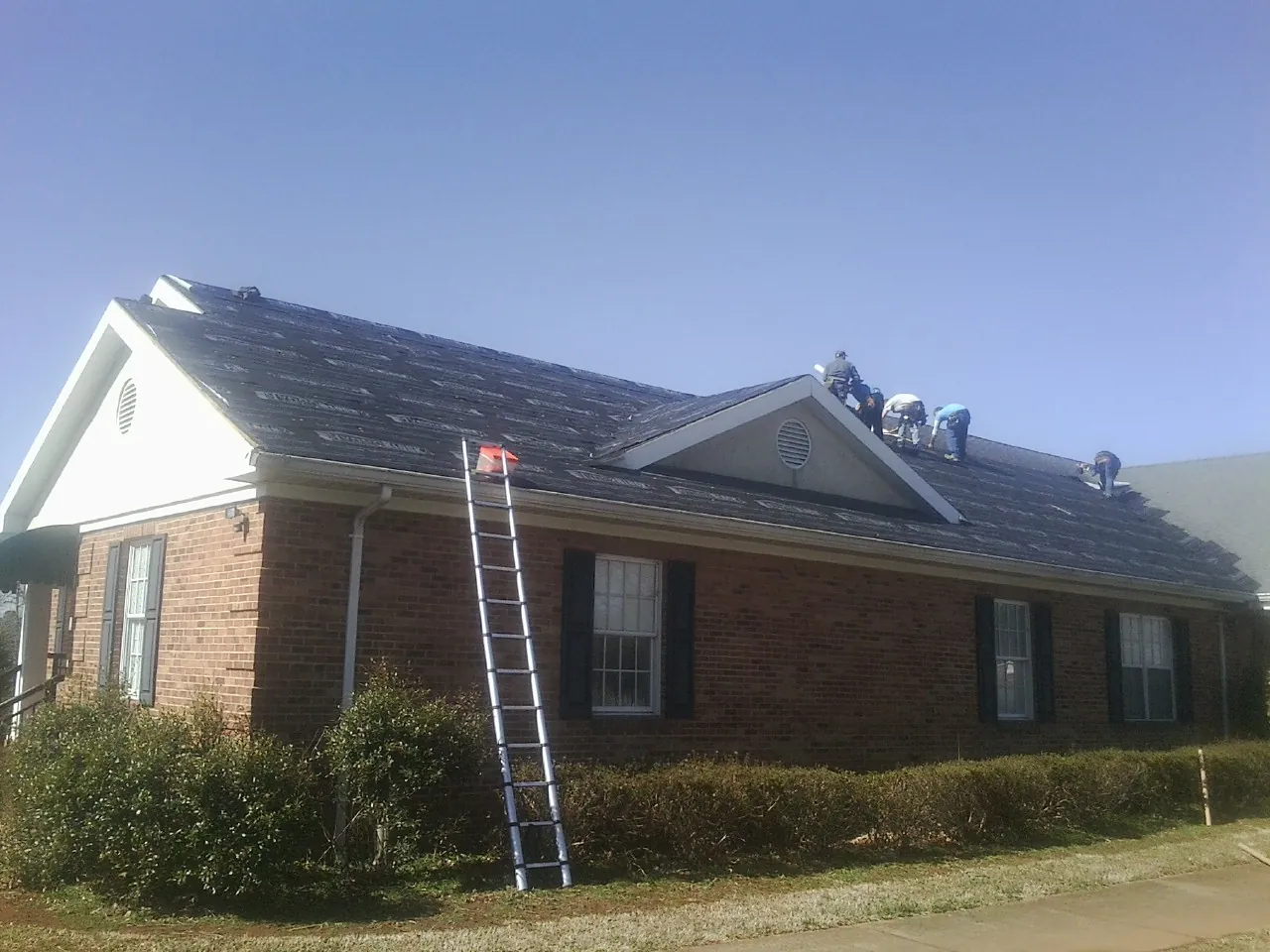 Crew tearing shingles off a brick ranch roof with a ladder set — Emergency Roof Repair work by our crew in Gibson