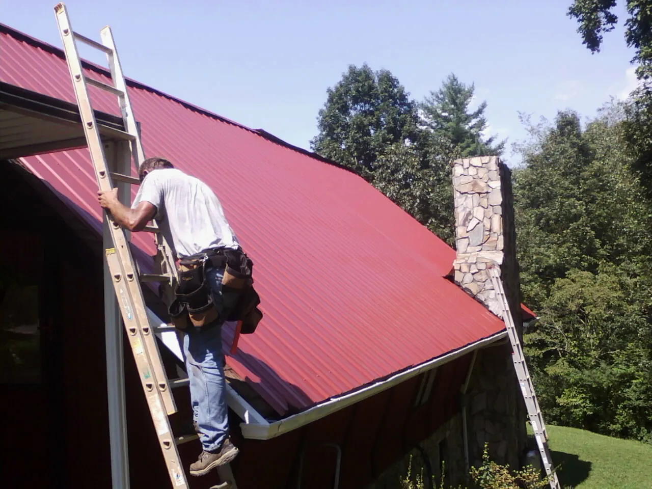 Roofer on a ladder beside a red metal roof and stone chimney — Soffit & Fascia Repair by Roofing Brothers in Gibson